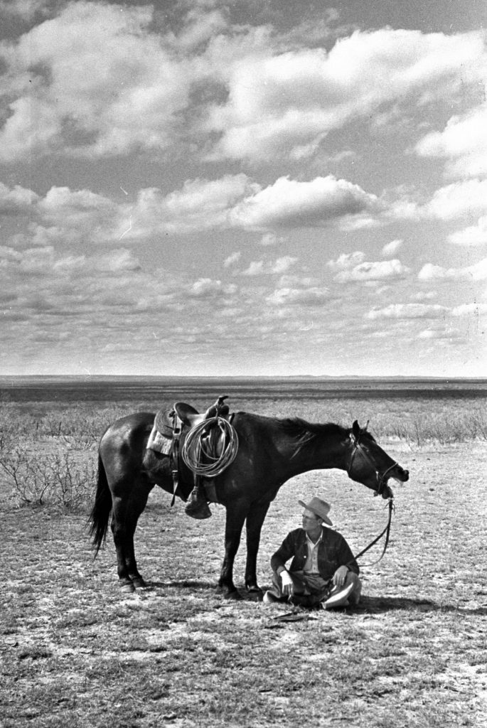 Foreman of the JA Ranch Clarence Hailey Long sitting in shade of his horse on prairie, 1949.